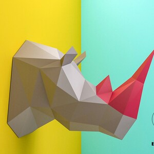 Pode incluir: Uma cabeça de rinoceronte de papel cinza com um chifre vermelho, montada em um fundo amarelo e azul. O rinoceronte é feito de papel dobrado e tem um design geométrico. O fundo é uma cor dividida, com amarelo à esquerda e azul à direita.