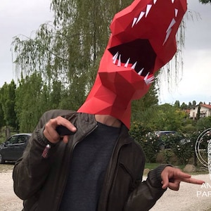 K&ouml;nnte beinhalten: Eine Person tr&auml;gt einen roten Papier-Dinosaurierkopf mit scharfen Z&auml;hnen und einem Blitz-Design. Die Person tr&auml;gt eine braune Lederjacke und zeigt mit der rechten Hand. Der Hintergrund ist ein verschwommenes Bild von B&auml;umen und einem Haus.
