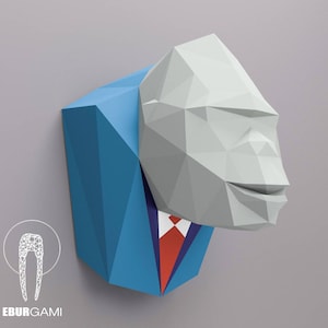 Pode incluir: Uma escultura geométrica em papel de uma cabeça de gorila usando um terno azul com uma gravata vermelha. A cabeça do gorila é cinza claro e o terno é azul vibrante. A escultura é montada em um fundo cinza. O logotipo "EBURGAMI" está no canto inferior esquerdo.