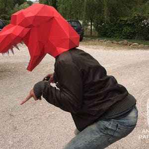K&ouml;nnte beinhalten: Eine Person tr&auml;gt eine rote Papier-Dinosauriermaske. Die Maske ist detailliert und hat scharfe Z&auml;hne. Die Person tr&auml;gt eine braune Jacke und eine blaue Jeans.
