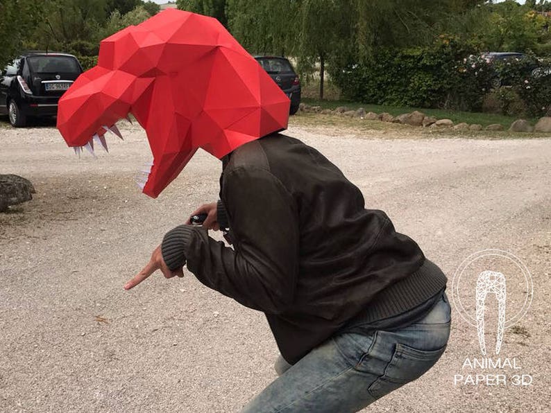 K&ouml;nnte beinhalten: Eine Person tr&auml;gt eine rote Papier-Dinosauriermaske. Die Maske ist detailliert und hat scharfe Z&auml;hne. Die Person tr&auml;gt eine braune Jacke und eine blaue Jeans.