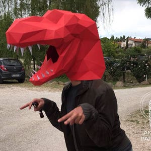 K&ouml;nnte beinhalten: Eine Person tr&auml;gt einen roten Papiermach&eacute;-Dinosaurierkopf. Der Dinosaurierkopf hat wei&szlig;e Z&auml;hne und ist aus gefaltetem Papier gefertigt. Die Person tr&auml;gt eine braune Jacke und schaut in die Kamera.