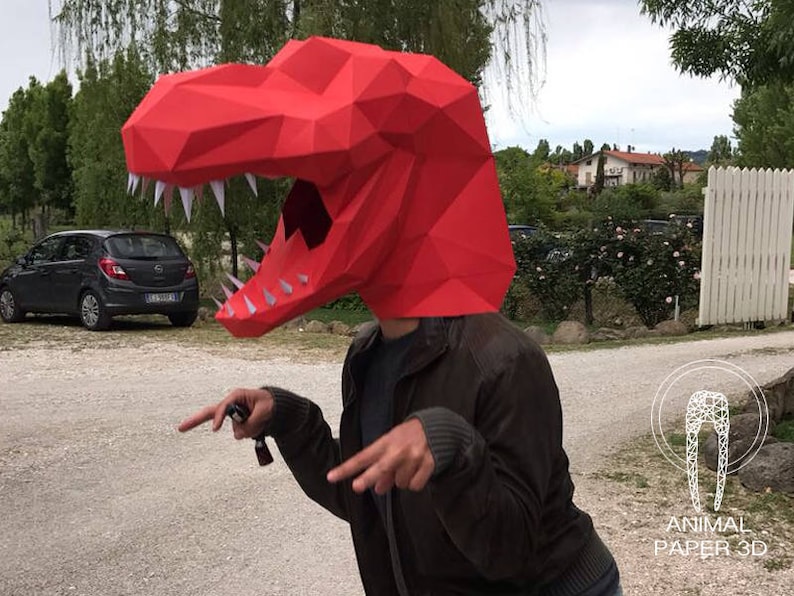 K&ouml;nnte beinhalten: Eine Person tr&auml;gt einen roten Papiermach&eacute;-Dinosaurierkopf. Der Dinosaurierkopf hat wei&szlig;e Z&auml;hne und ist aus gefaltetem Papier gefertigt. Die Person tr&auml;gt eine braune Jacke und schaut in die Kamera.