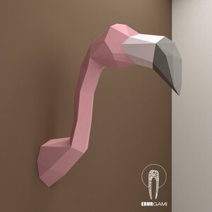 Pode incluir: Uma escultura de cabeça de flamingo rosa de papel com um bico cinza. A escultura está montada em uma parede marrom. O texto "EburGami" é visível na parte inferior da imagem.