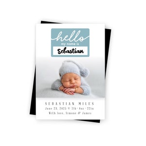 May include: A baby announcement card with a photo of a newborn baby boy wearing a gray knitted hat. The card has a blue and white name tag with the text "hello my name is Sebastian". The card also includes the baby's name, birthdate, weight, length, and the parents' names.