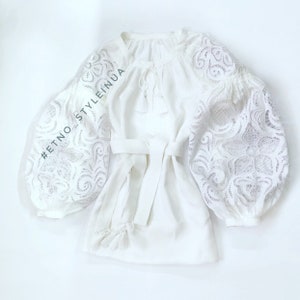 May include: White linen blouse with a lace design on the sleeves and a tie at the waist. The blouse has a hashtag #ETNO_STYLEINUA.