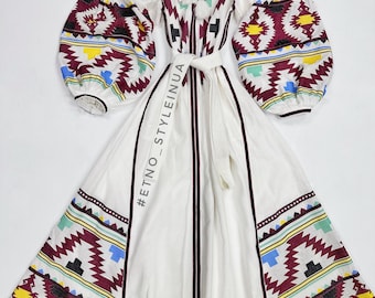 Puede incluir: Vestido blanco de lino con un patrón bordado geométrico en tonos de rojo, amarillo, azul y verde. El vestido tiene manga larga, cuello redondo y cinturón de lazo. #ETNO_STYLEINUA