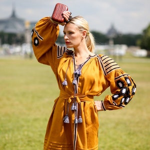 May include: A woman wearing a yellow embroidered dress with black and white details. She is holding a brown clutch purse and has her hair pulled back.