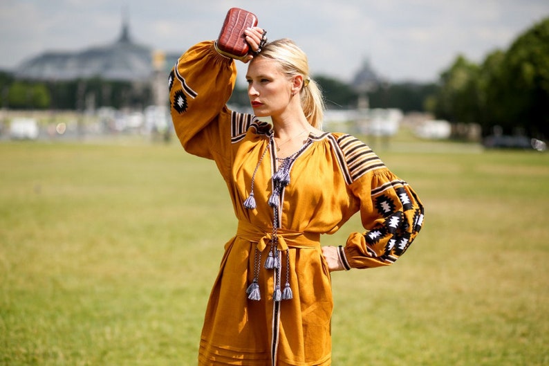 May include: A woman wearing a yellow embroidered dress with black and white details. She is holding a brown clutch purse and has her hair pulled back.