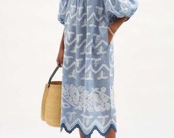 May include: A light blue midi dress with white embroidered floral and geometric patterns. The dress has a round neckline, puffed sleeves, and a scalloped hem. The model is wearing the dress with white sandals and carrying a straw tote bag.