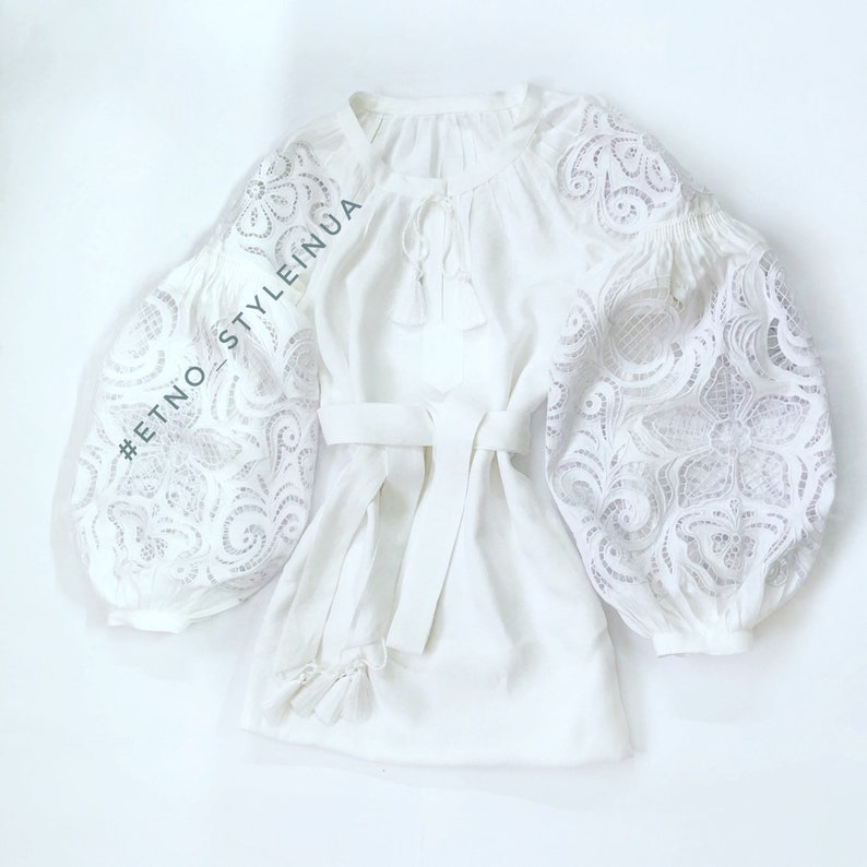 May include: White linen blouse with a lace design on the sleeves and a tie at the waist. The blouse has a hashtag #ETNO_STYLEINUA.