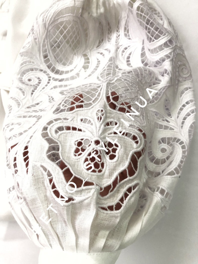 May include: White cotton fabric with a delicate lace pattern. The lace is cut out, creating a see-through effect. The fabric is gathered at the bottom, creating a ruffled look.
