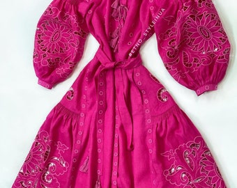 May include: A vibrant fuchsia dress with intricate floral cutouts on the sleeves and skirt. The dress features long, puffed sleeves, a tie waist, and decorative tassels. The design includes a series of small, evenly spaced circular cutouts and is made of a lightweight fabric.