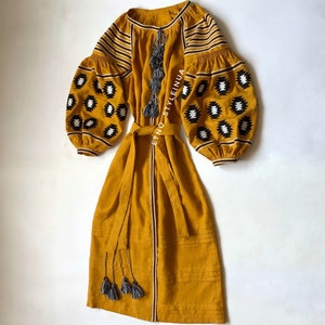 May include: A mustard yellow linen dress with black and white geometric embroidery on the sleeves and a matching fabric belt. The dress has a long, flowing skirt and a round neckline. The dress is accented with gray tassels.