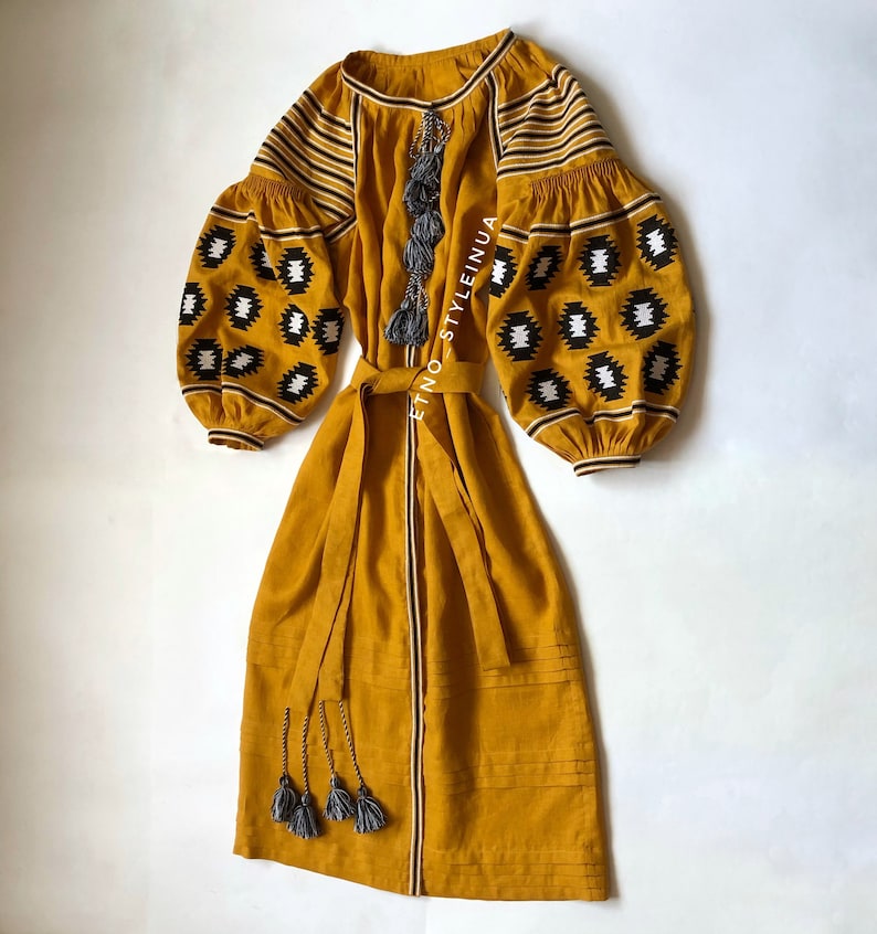 May include: A mustard yellow linen dress with black and white geometric embroidery on the sleeves and a matching fabric belt. The dress has a long, flowing skirt and a round neckline. The dress is accented with gray tassels.