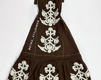 Puede incluir: Un vestido de lino marrón con diseños florales bordados en blanco. El vestido tiene un corpiño ajustado con escote en V y tirantes ajustables. La falda es amplia y fluida con un diseño de capas. El vestido tiene un ribete marrón alrededor del escote y el dobladillo. El vestido está etiquetado con el hashtag #ETNO_STYLEINUA.