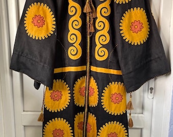 May include: Black dress with long sleeves, adorned with large yellow and orange floral patterns. The dress features a round neckline, decorative tassels, and yellow trim. The design includes a central vertical pattern of yellow swirls.