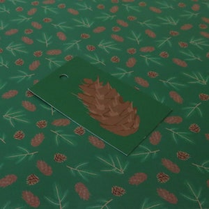 May include: A green gift tag with a brown pine cone illustration. The tag has a hole punched in the top for string or ribbon.
