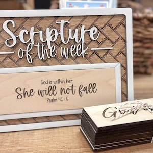 May include: Wooden framed sign with the text "Scripture of the week" and "God is within her, She will not fall, Psalm 46:5". A stack of wooden cards with the word "God" is also visible. The frame has a white border.