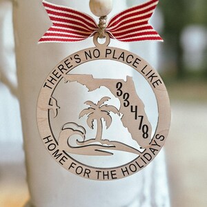 May include: Wooden Christmas ornament with a Florida state outline, a palm tree, and the text "THERE'S NO PLACE LIKE HOME FOR THE HOLIDAYS" and "33418". The ornament is decorated with a red and white striped bow and a wooden bead.