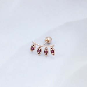 May include: A gold ear cuff with a curved bar and five small, red gemstone accents. The gemstones are set in a leaf-like shape.