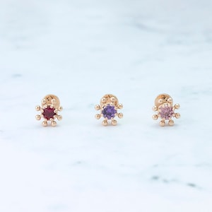 May include: Three gold-toned earrings with a starburst design and a single gemstone in the center. The earrings feature a red, purple, and pink gemstone.