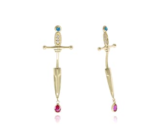 May include: A pair of gold earrings with a sword design. Each earring features a blue gemstone at the top of the sword and a red gemstone dangling from the bottom.