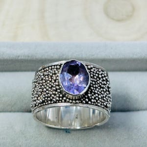 May include: A wide silver ring featuring a large, oval, purple gemstone. The band is textured with small, round silver beads. The ring is displayed on a light grey surface.