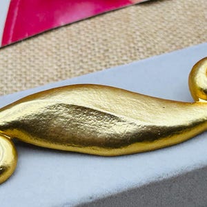 Puede incluir: Broche dorado con un diseño en espiral en cada extremo. El broche tiene una superficie texturizada y una forma curva, similar a una hoja. Descansa sobre una superficie gris, lo que sugiere una joya o accesorio.