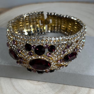 Pode incluir: Uma pulseira larga de punho articulado com base de metal dourado, incrustada com strass brancos transparentes, cristais mais pequenos de cor violeta claro e várias pedras grandes ovais de cor violeta escuro facetadas em padrão.