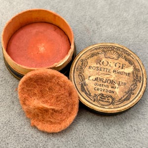 Könnte beinhalten: Eine antike runde Dose Rouge mit schwarzem Deckel und einem Etikett mit der Aufschrift "Rouge Rosette Prume L'Jurjois Ltd Queens Way Croydon Made in England". Die Dose ist geöffnet und das Rouge ist leuchtend orange.