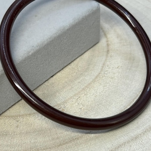 May include: A dark brown, oval bangle bracelet. The smooth, polished surface reflects light, highlighting its circular form. The bracelet is resting on a light grey surface and a wooden background.