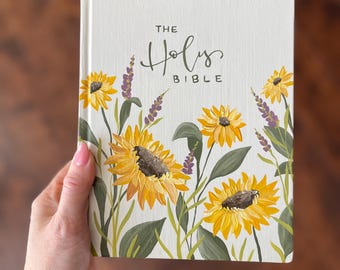 May include: A white hardcover book with the words "The Holy Bible" printed on the front cover. The cover is decorated with a floral pattern of sunflowers and purple flowers.