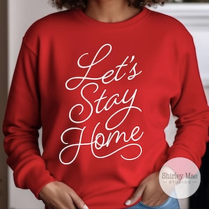 Puede incluir: Sudadera roja con texto blanco que dice "Let's Stay Home". El texto está en una fuente cursiva. La sudadera tiene un pequeño logotipo en la esquina inferior derecha que dice "Shirley Mae Studios".