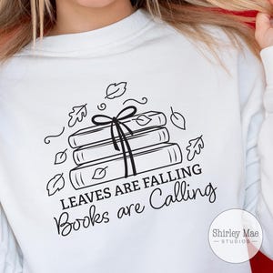 Puede incluir: Sudadera blanca con un diseño gráfico en blanco y negro de una pila de libros con un lazo en la parte superior. El texto "Leaves are falling, Books are calling" está debajo del gráfico. El texto "Shirley Mae Studios" está en la esquina inferior derecha.
