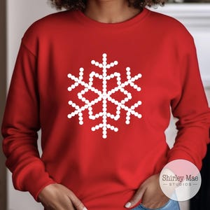 May include: Red sweatshirt with a white snowflake design. The snowflake is made up of small white circles. The sweatshirt has the text "Shirley Mae Studios" on the bottom right corner.
