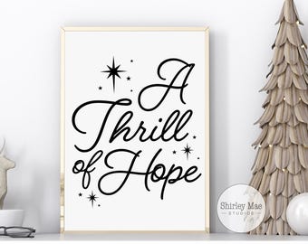 May include: A black and white printable art print with the text "A Thrill of Hope" in a decorative font. The text is surrounded by small stars. The print is framed and hanging on a white wall.