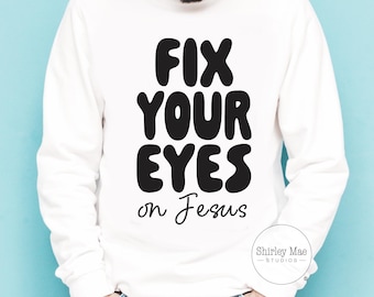 May include: White long-sleeve sweatshirt with the text "FIX YOUR EYES on Jesus" in black. The sweatshirt also features a small circular logo that says "Shirley Mae Studios".