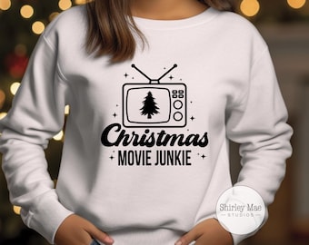 May include: White sweatshirt with a black graphic of a retro television with a Christmas tree on the screen. The text "Christmas Movie Junkie" is below the television graphic.