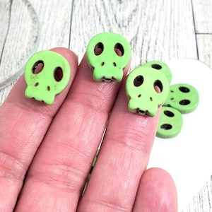 May include: Close-up of several small, light green skull-shaped beads. The beads have black eye and nose sockets. Some beads are in focus, while others are slightly blurred. The beads are on a white surface.