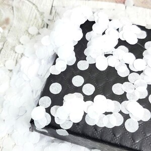 May include: A black gift box with white polka dots is covered in white confetti. The box has a quilted pattern and is sitting on a light-colored wooden surface. The confetti is scattered around the box.