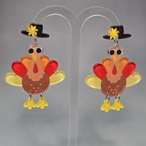 May include: A pair of dangle earrings featuring a turkey design. Each turkey has a brown body, yellow feet, and a multi-colored tail with red, orange, and yellow translucent acrylic pieces. A black hat with a yellow star sits atop each turkey head.