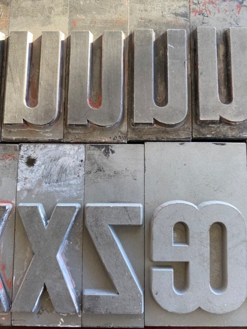 Pu&ograve; includere: Primo piano di blocchi di stampa tipografica in metallo, che mostrano le lettere 'U', 'W', 'X', 'Z' e '90'. I blocchi sono grigi e hanno una superficie usurata e testurizzata.