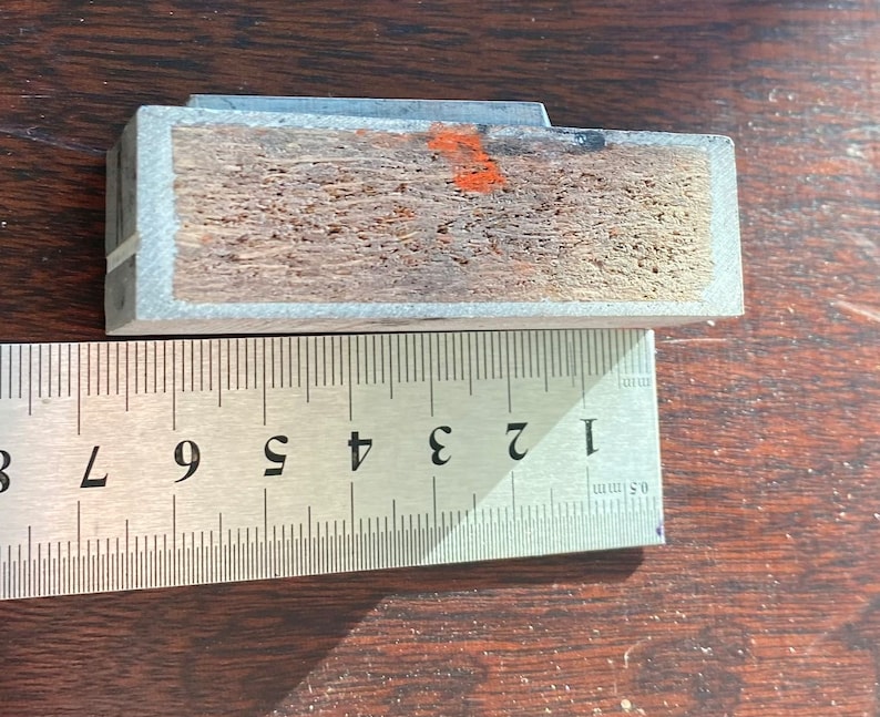 Pu&ograve; includere: Primo piano di un blocco di legno con una cornice in metallo grigio. Il blocco &egrave; largo circa 2,5 cm e lungo 7,5 cm. Il legno ha una venatura naturale ed &egrave; leggermente rovinato.