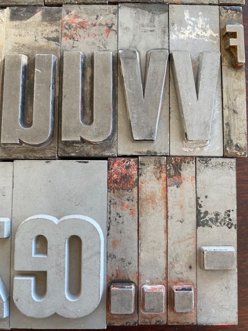 Pu&ograve; includere: Un set di blocchi di stampa tipografica in metallo vintage, tra cui le lettere U, V ed F, nonch&eacute; il numero 90. I blocchi sono in metallo grigio con un po' di ruggine e usura.