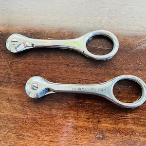 Puede incluir: Dos herramientas de metal plateado con un bucle en un extremo y un extremo redondeado en el otro. Las herramientas están sobre una superficie de madera.