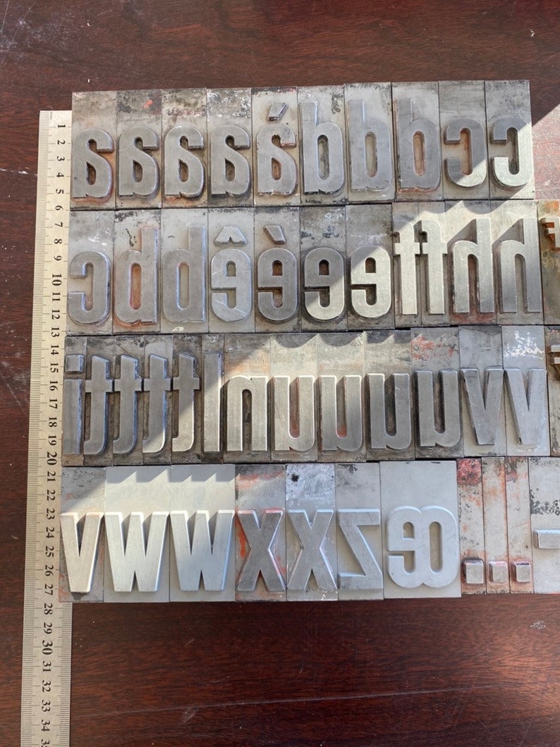 Pu&ograve; includere: Un set di blocchi di stampa tipografica in metallo vintage, che include le lettere c, d, e, f, g, h, i, j, l, m, n, o, p, q, r, s, t, u, v, w, x, y, z e il numero 0. I blocchi sono disposti in file e sono in metallo argentato.