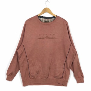 May include: A dusty rose sweatshirt with the text "SISSY by KANSAI YAMAMOTO" in brown. The sweatshirt has a crew neck with a plaid lining and a pocket on the front. Black trim accents the front and sleeves.
