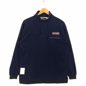 May include: A navy blue long-sleeve pullover shirt with a mock neck collar and a partial front placket. The chest features a rectangular patch with the text HILFIGER SAILING GEAR and a small horizontal zippered pocket. A white square tag is on the hem.