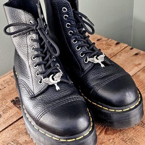 May include: A pair of black leather platform boots with silver-tone moth charms on the laces. The boots have a textured finish, yellow stitching, and black laces. The boots are on a wooden surface.
